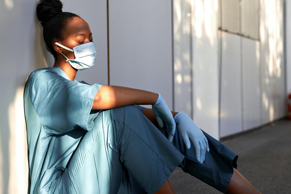 Tired exhausted female african scrub nurse wears face mask blue uniform gloves sits on hospital floor.